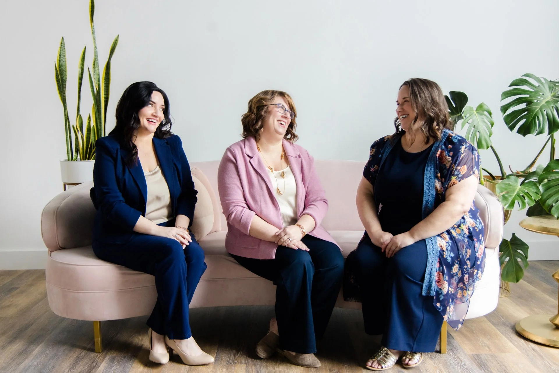 Three women sitting on a light pink sofa, smiling and talking together. They are in a room with green plant decorations and wooden flooring.