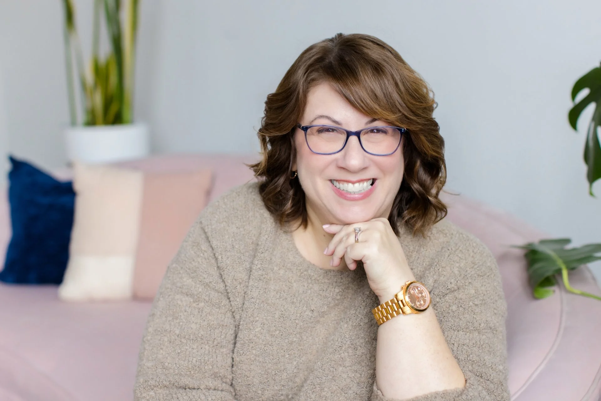 A woman with glasses, short brown hair, and a beige sweater smiling with her chin resting on her hand, sitting on a pink couch with pillows and plants in the background.