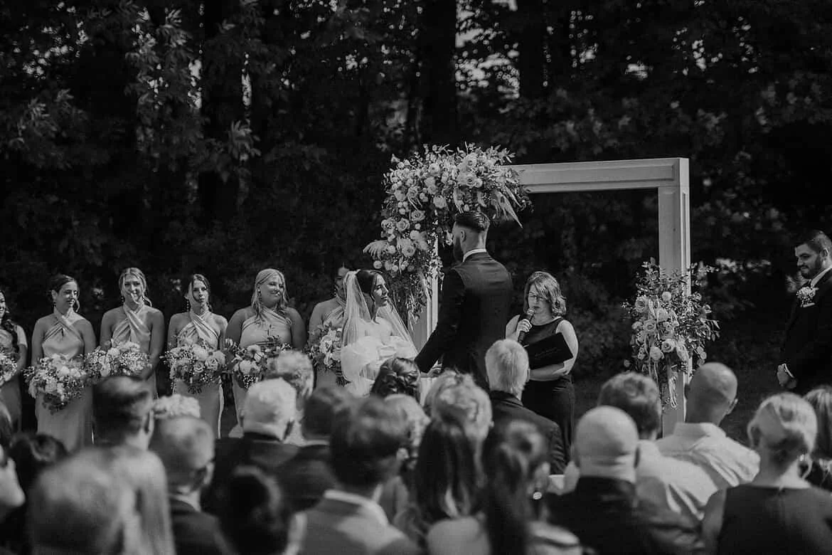 A black and white photo of an outdoor wedding ceremony with a couple at the altar, surrounded by bridesmaids and groomsmen, with guests seated in front. The altar is decorated with floral arrangements and a large arch.