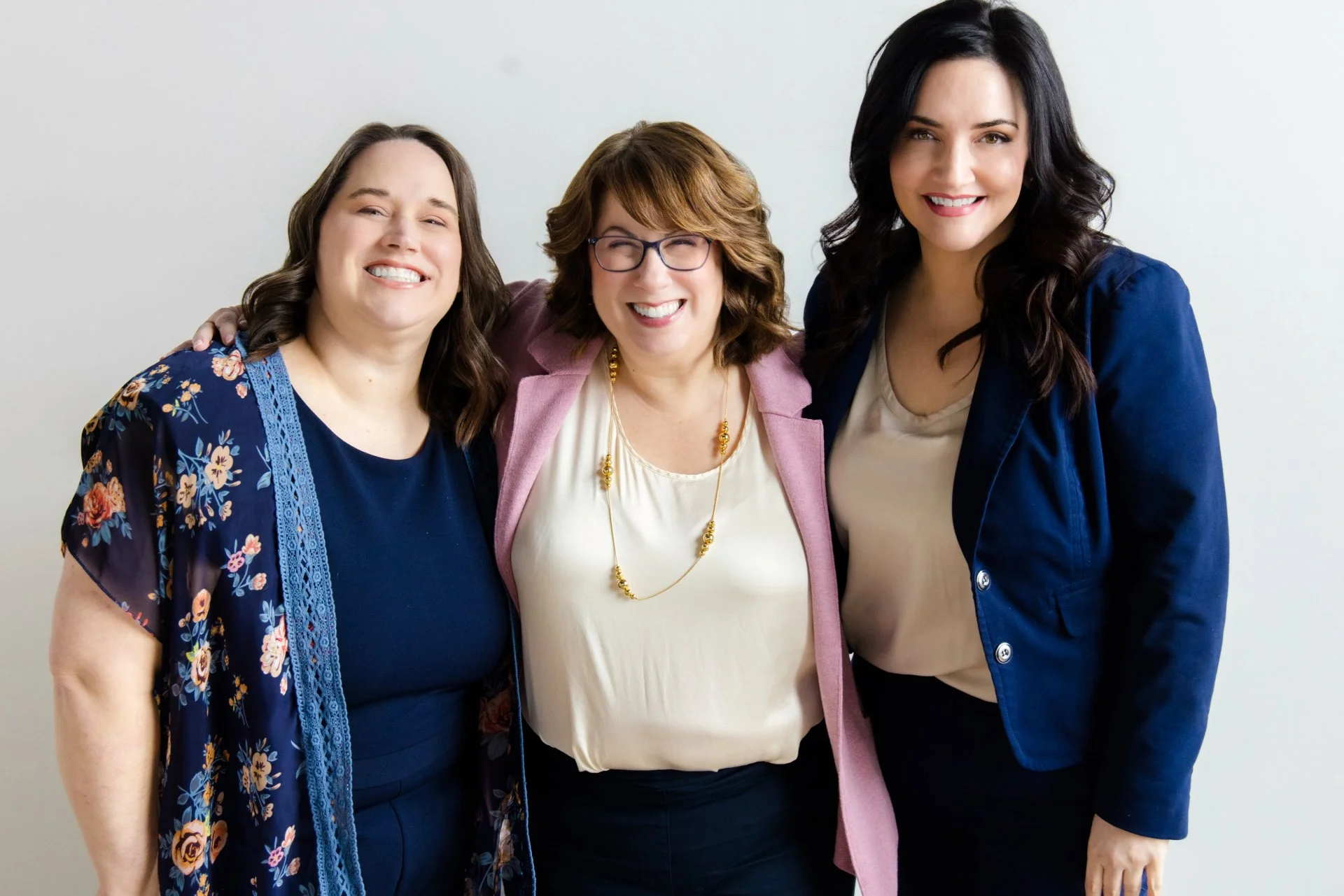 Three women standing close together, smiling, with a plain white background.