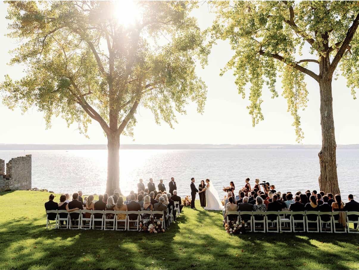 Outdoor wedding ceremony by a lake with guests seated on white chairs, two large trees, and the sun shining through the branches.