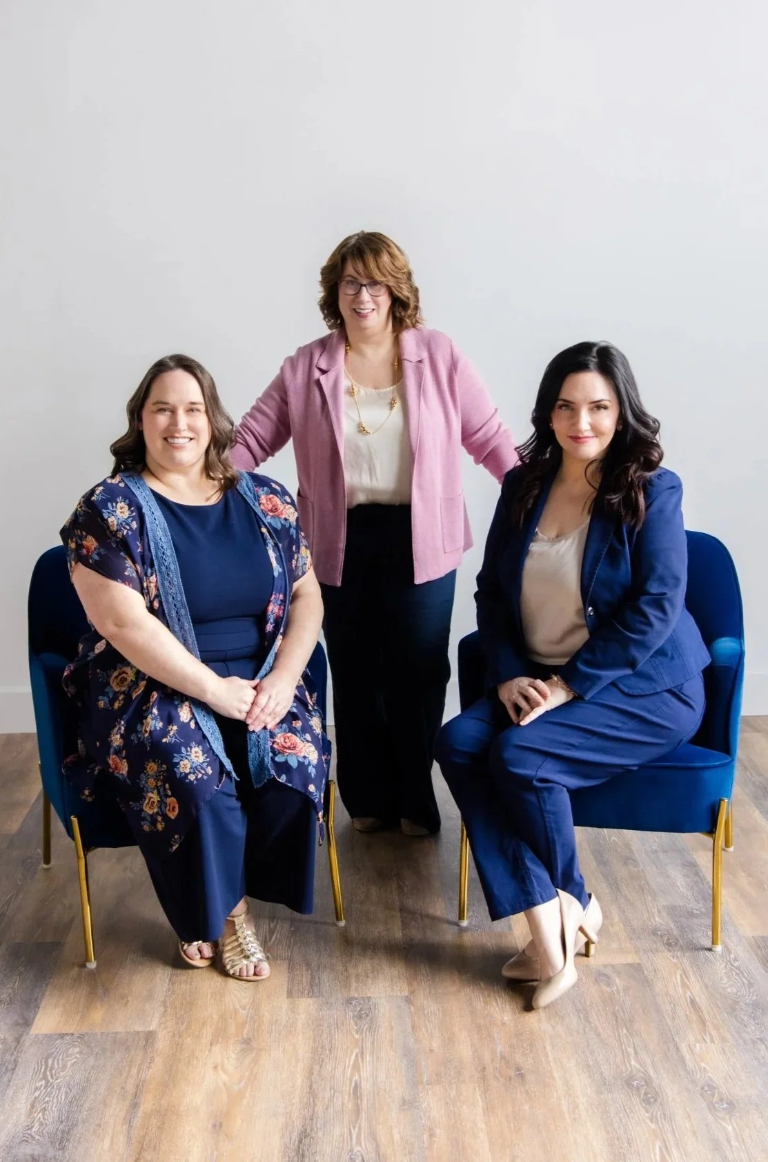 Three women in professional attire, two sitting in blue chairs and one standing behind them, posing for a photo in a minimalistic room with wooden flooring and white wall background.