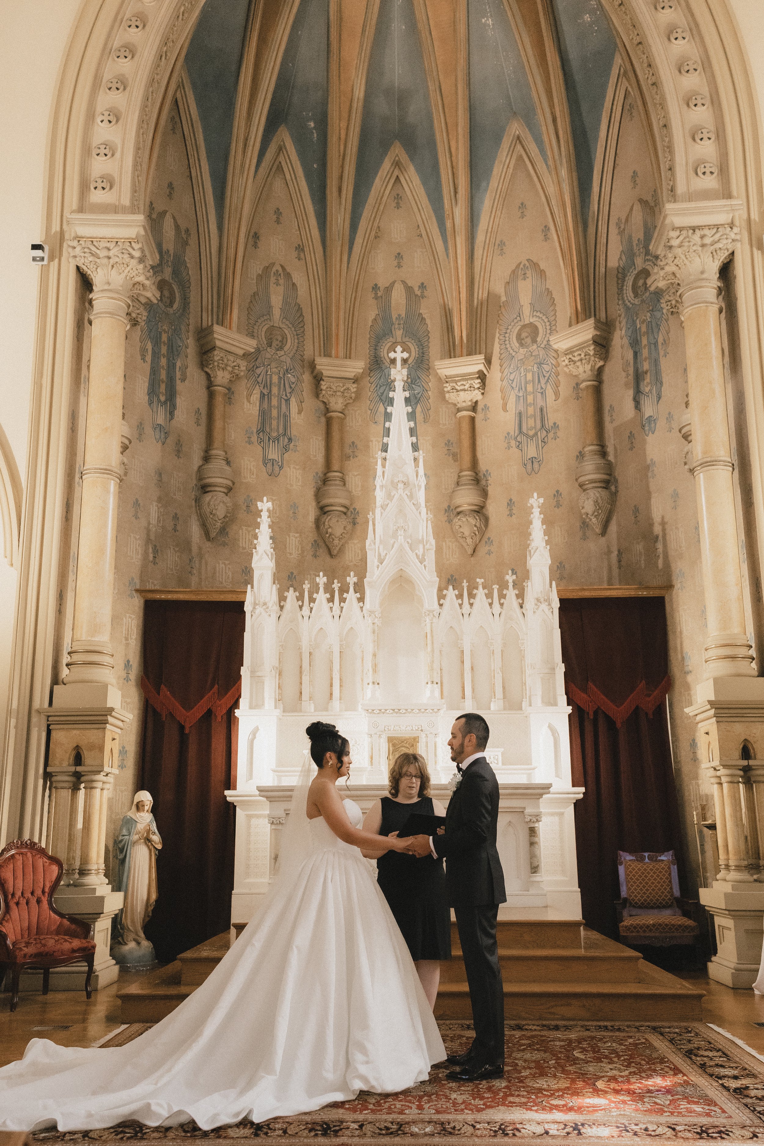 A bride and groom exchanging vows during a wedding ceremony inside a church, with a priest or officiant in the middle, surrounded by religious statues and ornate architectural details.