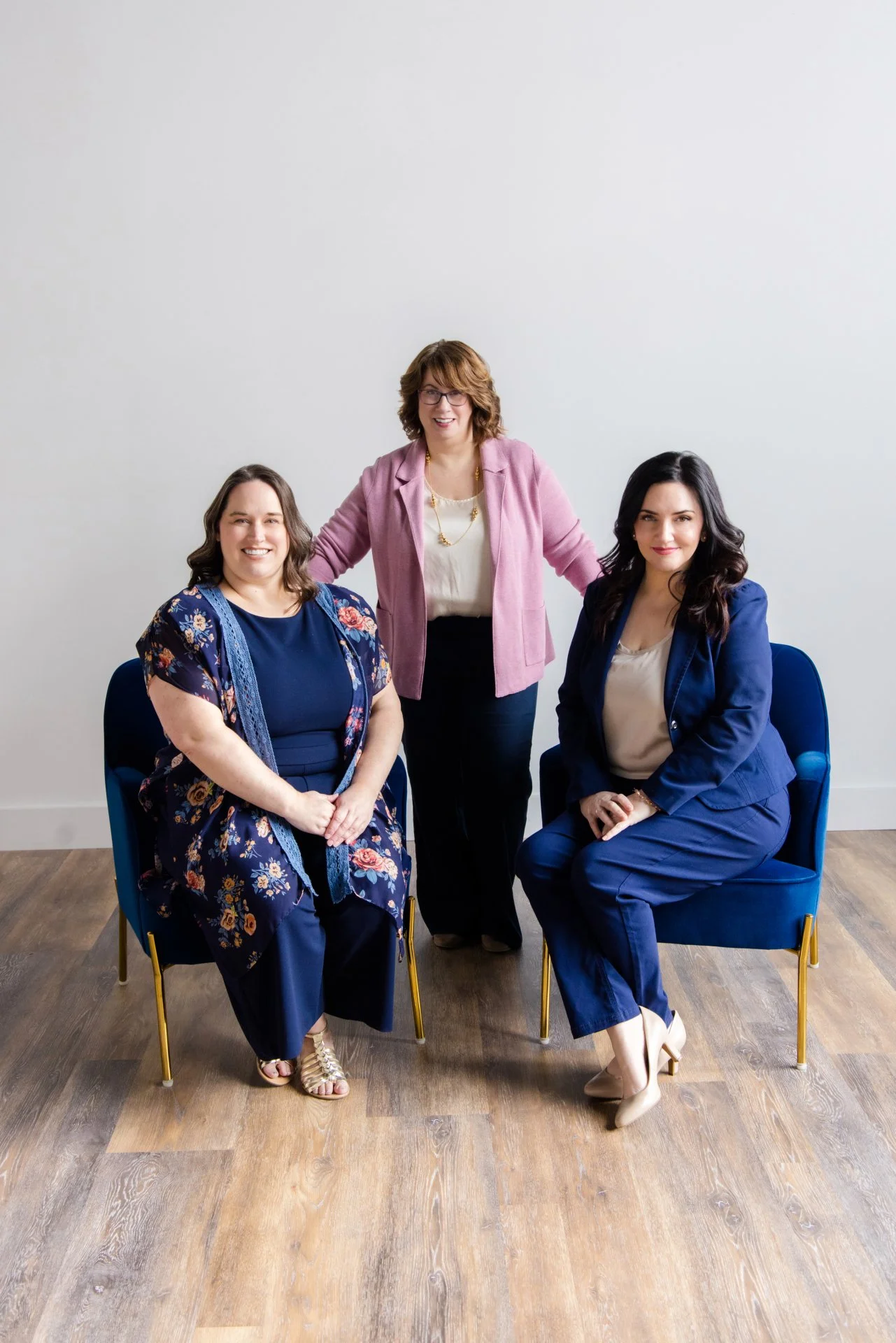 Two women sitting in navy blue chairs the one on the right in a blue suit, the one on the left is in a blue romper with a kimono over it.  There is a third woman standing behind them in a pink jacket, cream shirt, and n