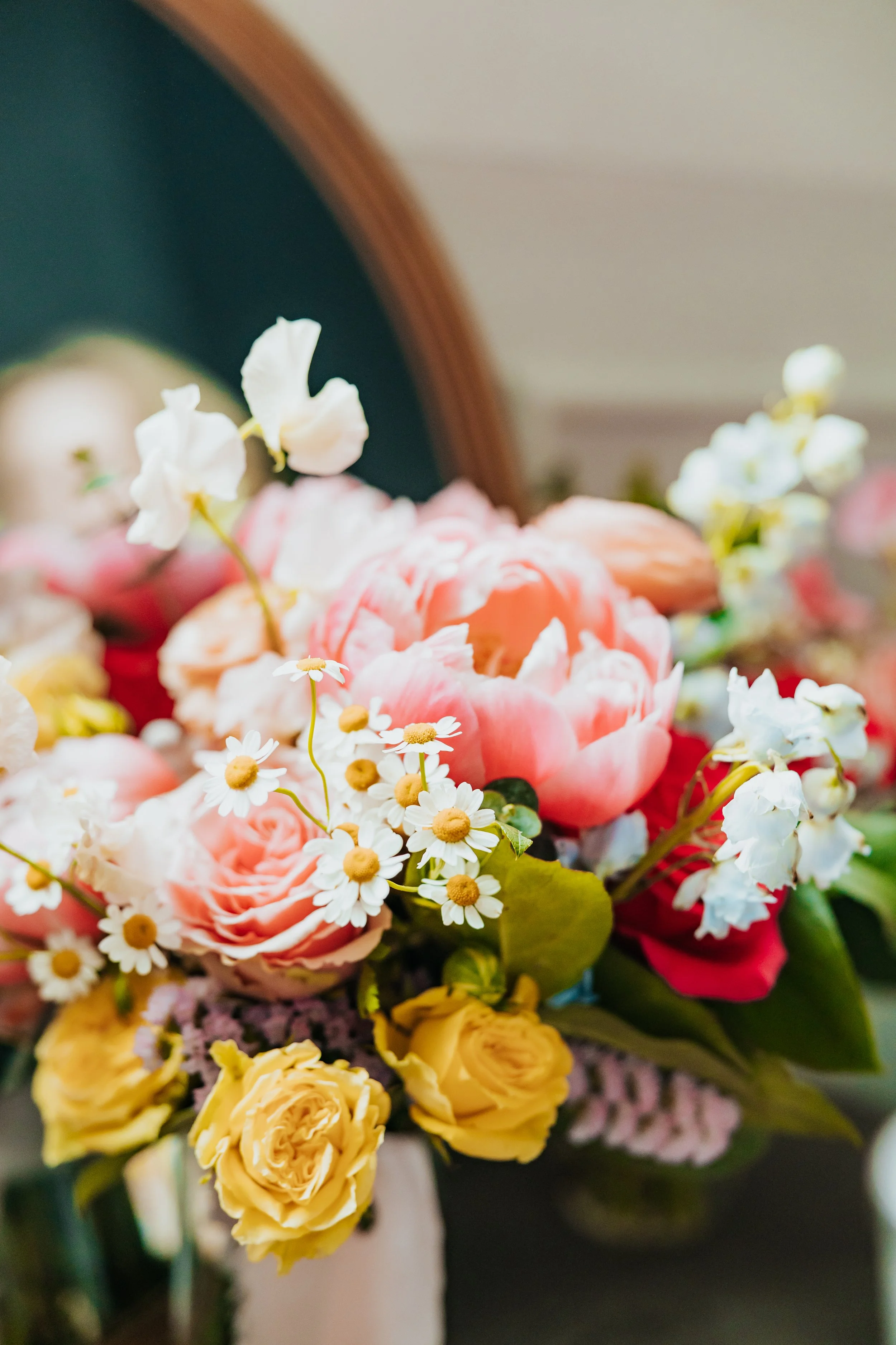Close-up of a colorful bouquet of flowers including pink roses, white daisies, white sweet peas, and yellow roses, reflected in an oval mirror.