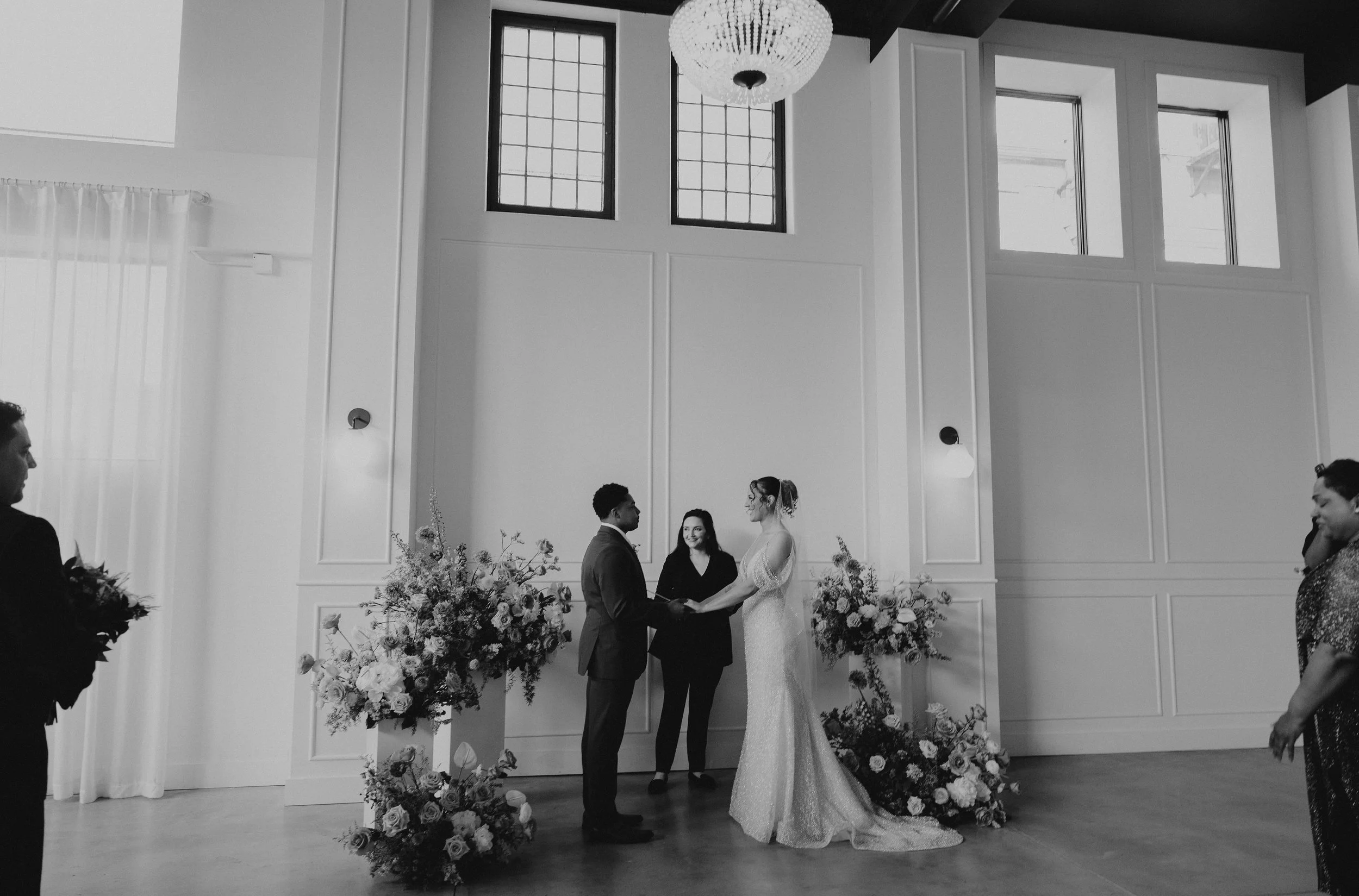 Black and white photo of a wedding ceremony with a bride and groom holding hands, standing in front of an officiant, inside a bright room with large windows, decorative wall paneling, and floral arrangements.