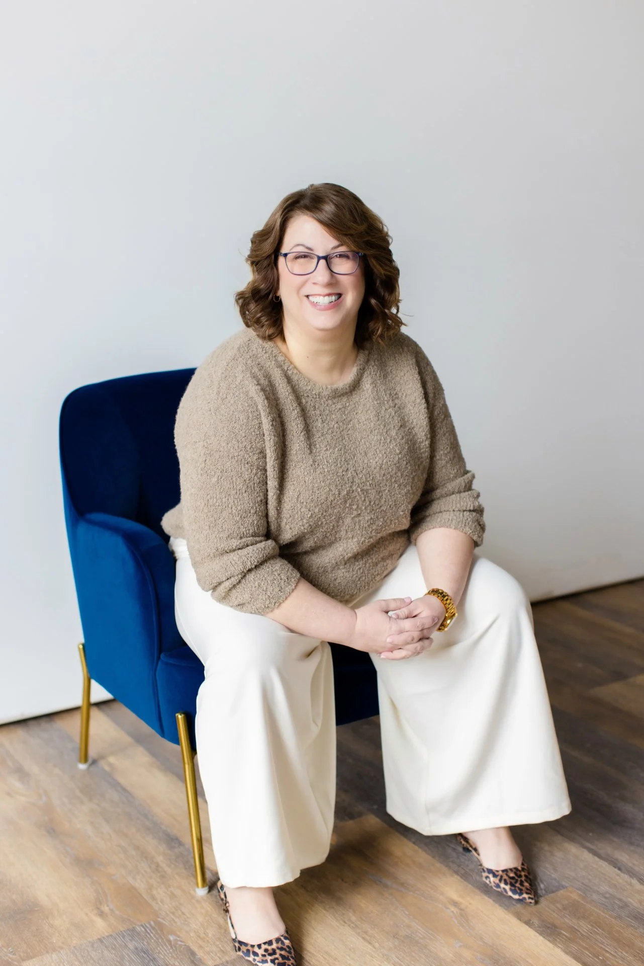 A woman with shoulder-length brown hair, wearing glasses, a beige sweater, white wide-leg pants, animal-print heels, and a gold bracelet, sitting and smiling on a blue armchair.