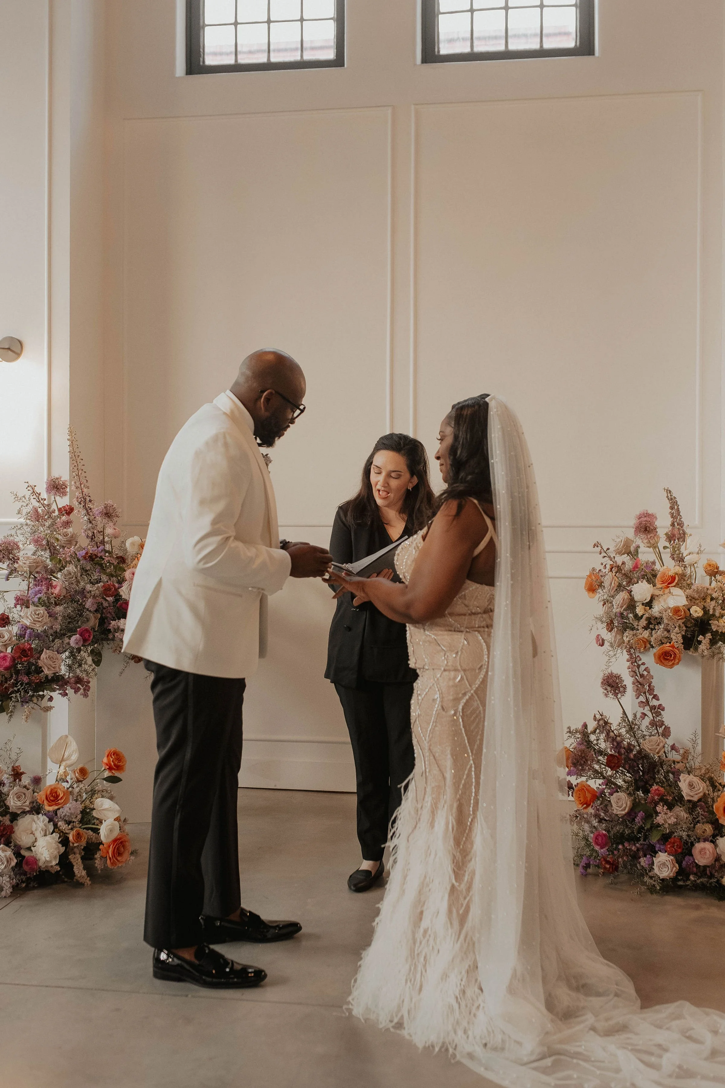 A woman in a black suit officiating a wedding for a woman in a wedding gown and and man in a tuxedo with the white jacket