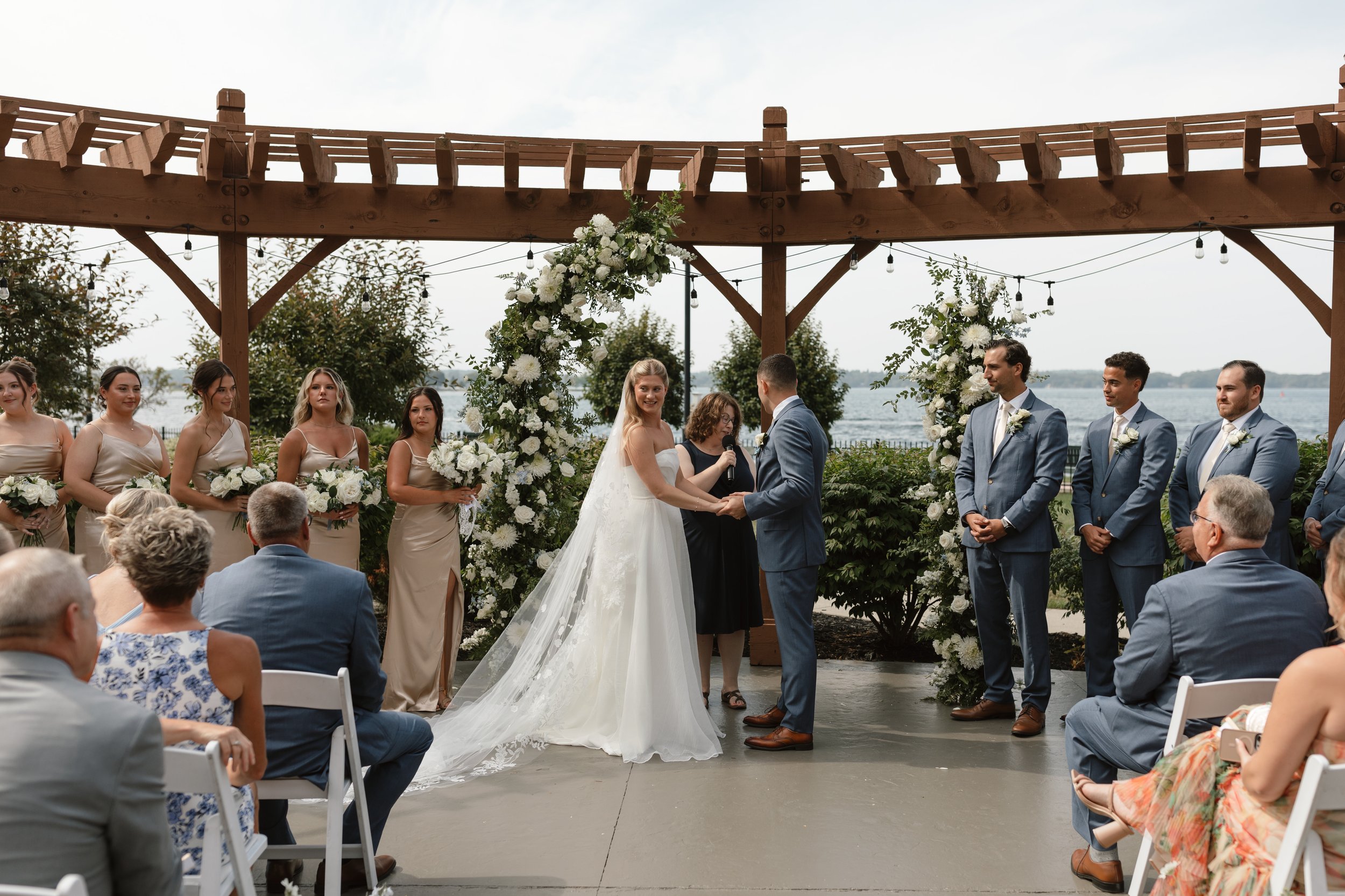 Wedding ceremony outdoors by a lake with a bride and groom holding hands, surrounded by bridesmaids and groomsmen, under a wooden arch decorated with white flowers and greenery.