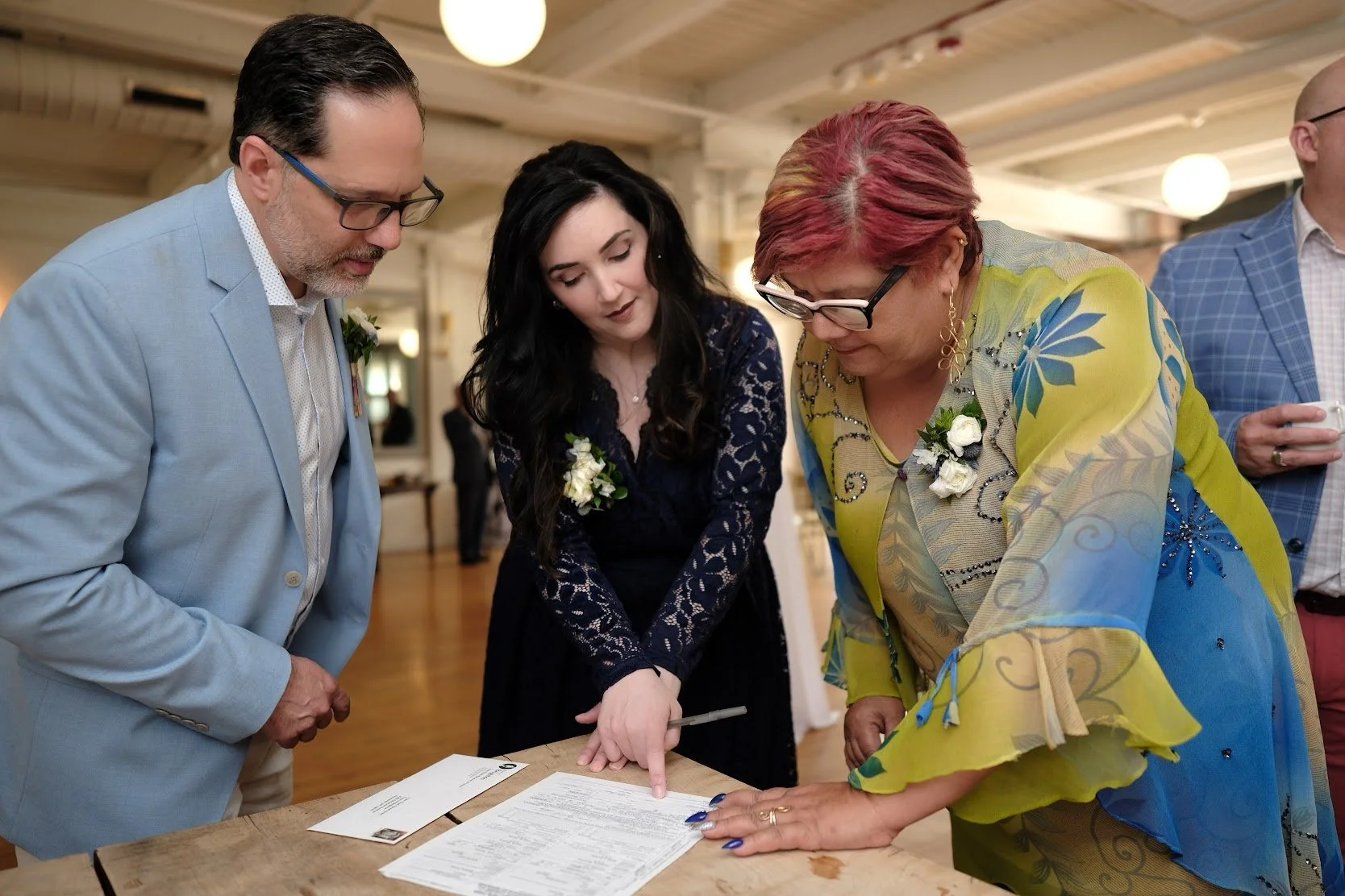Three adults are gathered around a table, looking at documents. They are dressed in formal attire, with two women wearing corsages. The setting appears to be a social or formal event in a warmly lit room.