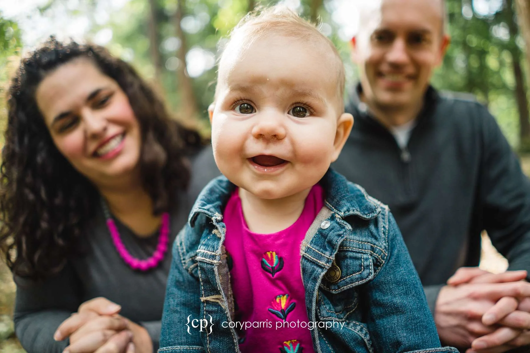 A little girl smiling for the camera in front of her parents at their family portrait session at the Washington Park Arboretum.
