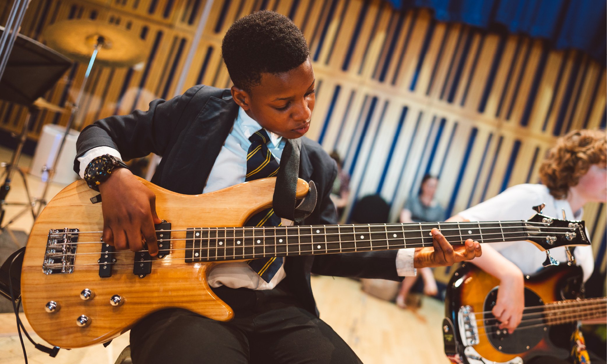 A young electric bass player wearing school uniform and holding a light brown bass guitar.