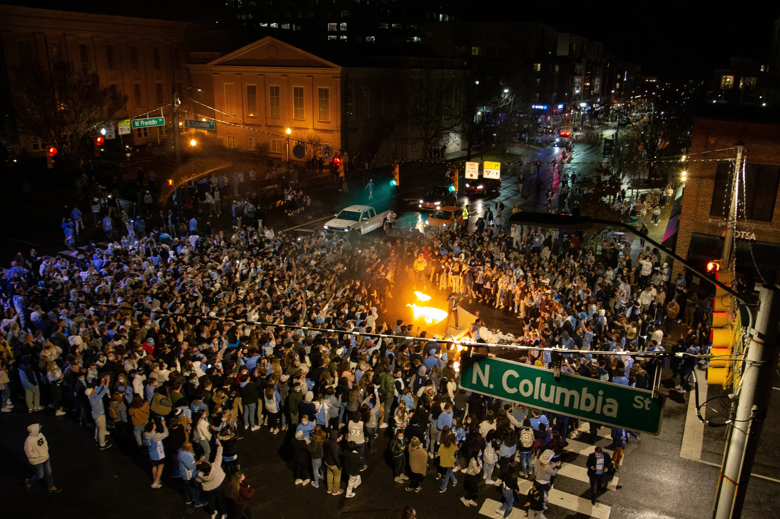 A large crowd of people gathered on a city street at night, with some holding torches or fire. Streetlights and city buildings are visible in the background, along with multiple cars and traffic signals.