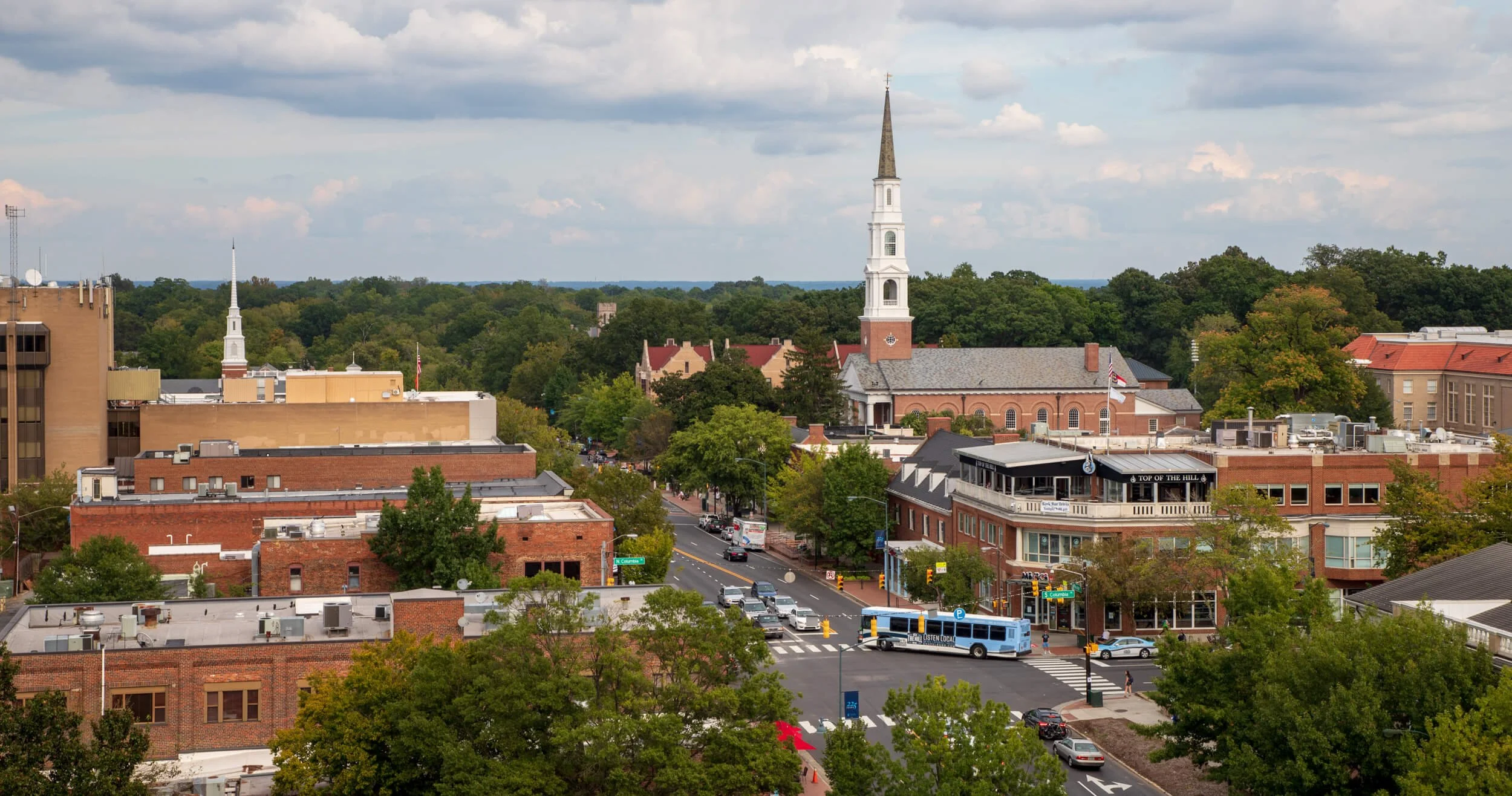 View of a small downtown area with brick buildings, a church with a tall steeple, a street with cars and a bus, trees, and a partly cloudy sky.