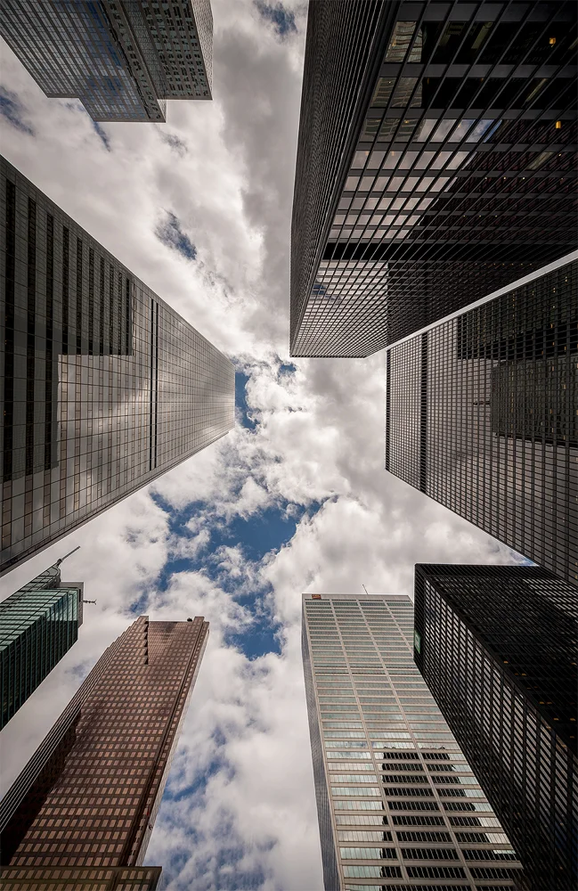 This photo is taken at around noon in downtown Toronto in the Financial District. Due to the time of day, the light at the ground level was not that special, so I started searching something interesting perspective-wise. After some walking around, I found a place from where the tall buildings looked nice when looking directly up to the sky. I waited a while in the same spot, letting the clouds pass and experimented with different-looking skies. I ended up liking a variant where there was a break in the clouds in between the buildings. The final image you see here is a three-exposure HDR post-processed using Lightroom and Photoshop.