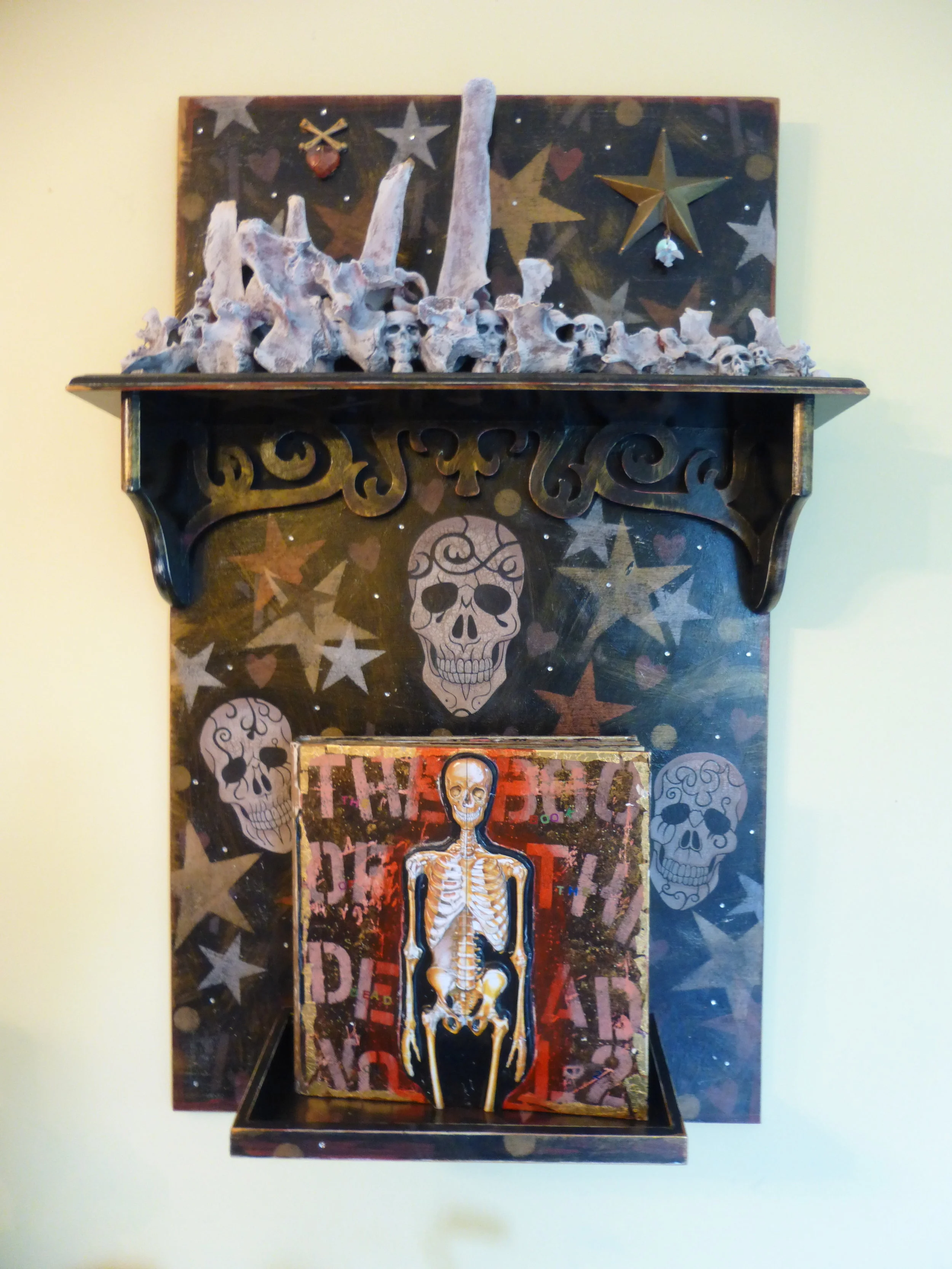 "the book of the dead - volume 2", mixed-media collage. Wood, moose vertebrae, plastic skulls, skeleton from the dollar store, acrylic paint, book, text.