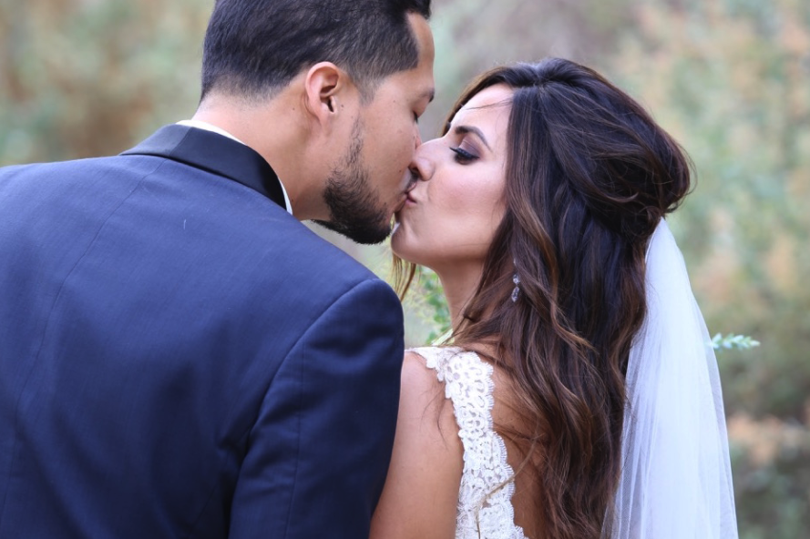 Brunette Bride with Veil and Hair down Kissing Husband in outdoor wedding photos. Bridal hair and makeup by Vanity Belle in Orange County (Costa Mesa) and San Diego (La Jolla)