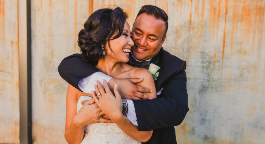 Asian Bride with Updo hugging husband smiling in Wedding Photos Outside. Bridal Hair and Makeup by Vanity Belle in Orange County (Costa Mesa) and San Diego (La Jolla)