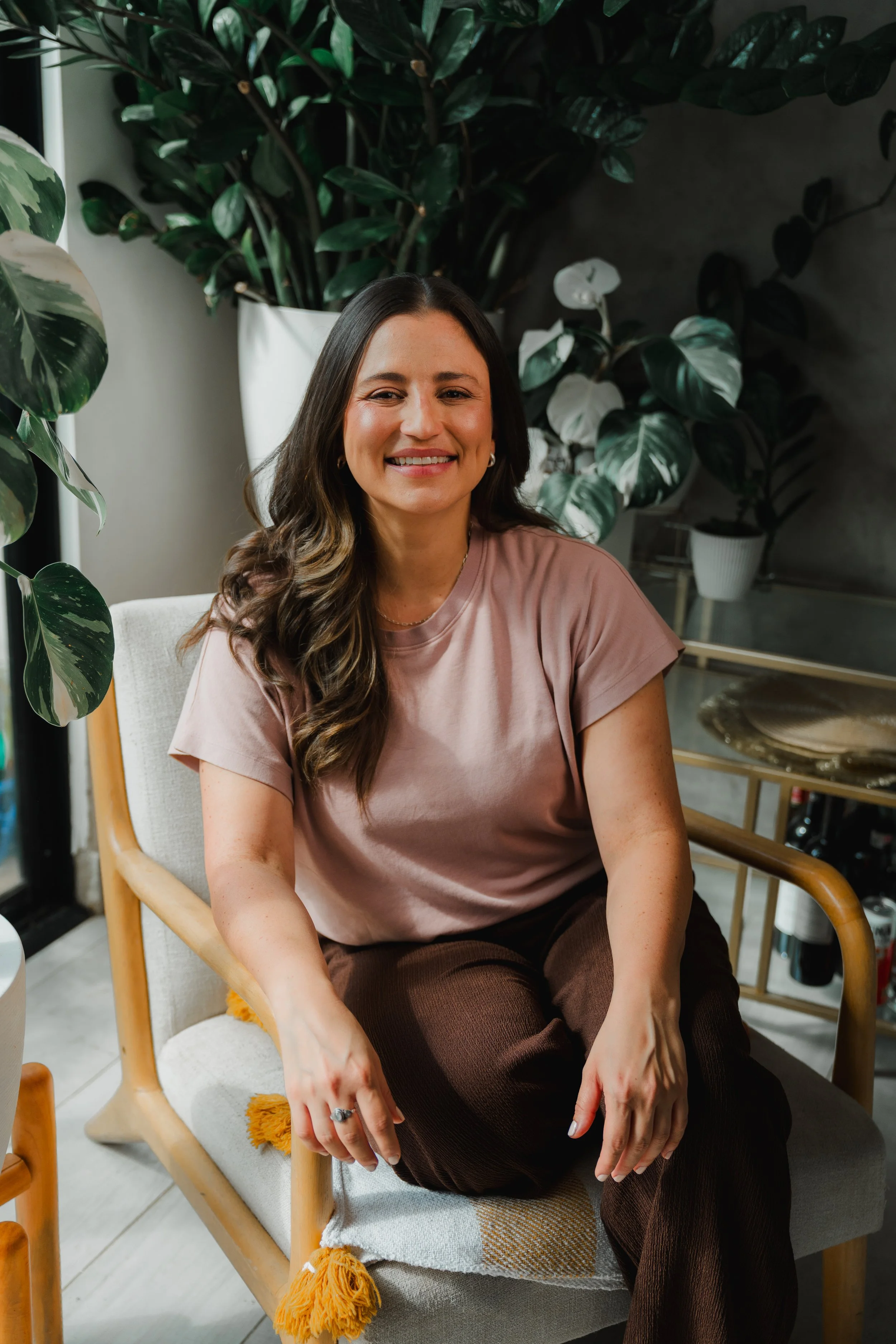 Mujer sonriendo, sentada en una sala con plantas verdes de fondo.