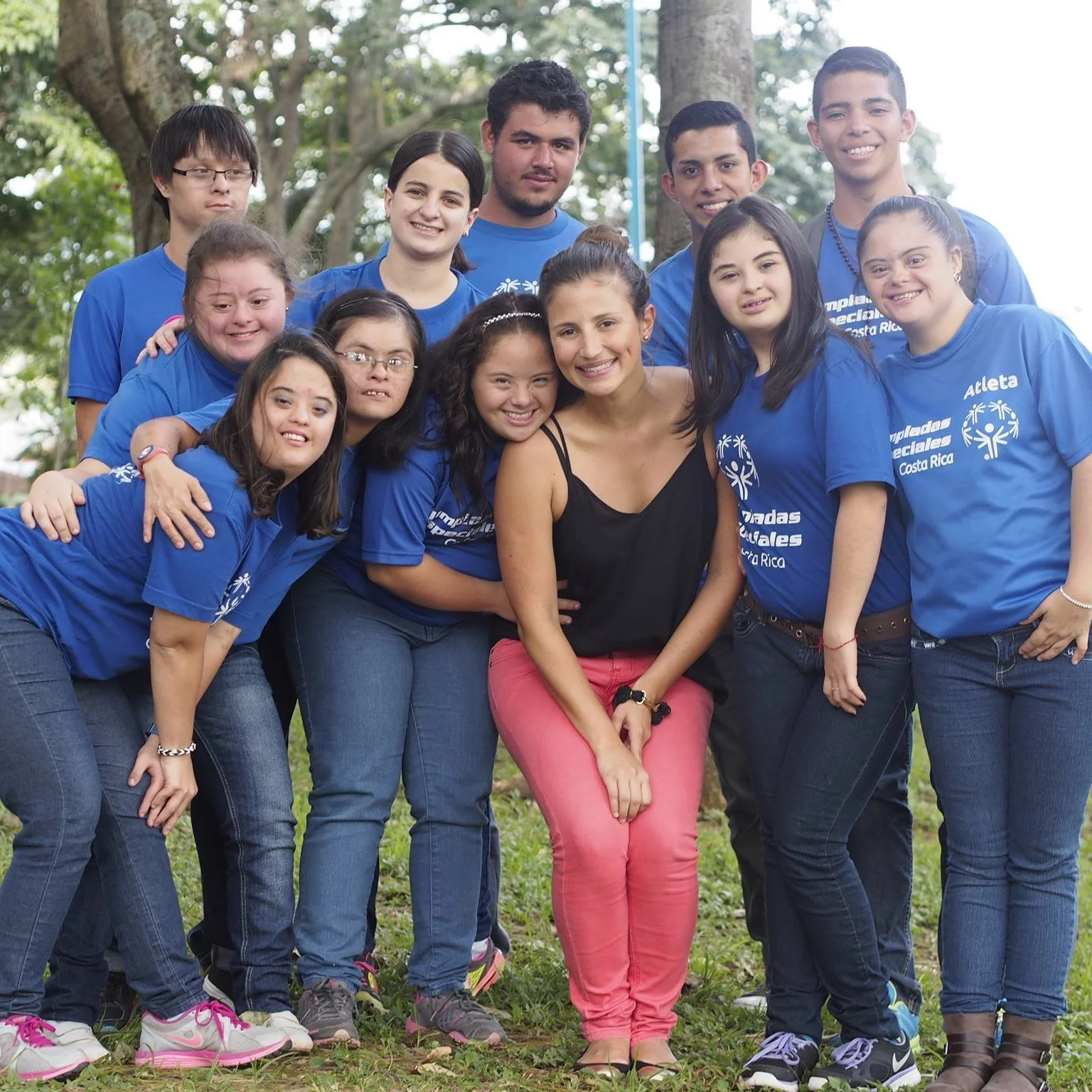 Grupo de personas jóvenes y una mujer adulta en un parque, algunos con camisetas azules de eventos deportivos en Costa Rica, sonriendo y posando para una foto grupal.