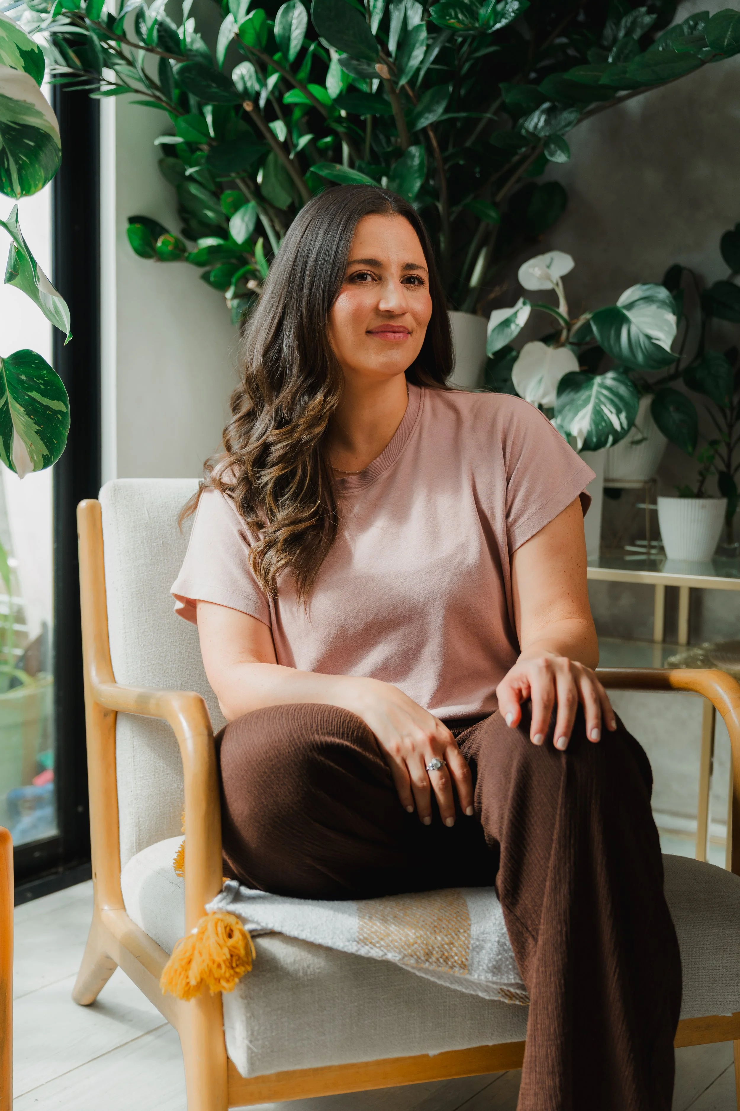 Una mujer con cabello castaño y rizado, vestida con camiseta rosa y pantalones marrón, sentada en una silla de madera y tela clara, rodeada de plantas verdes grandes, en un ambiente interior bien iluminado.