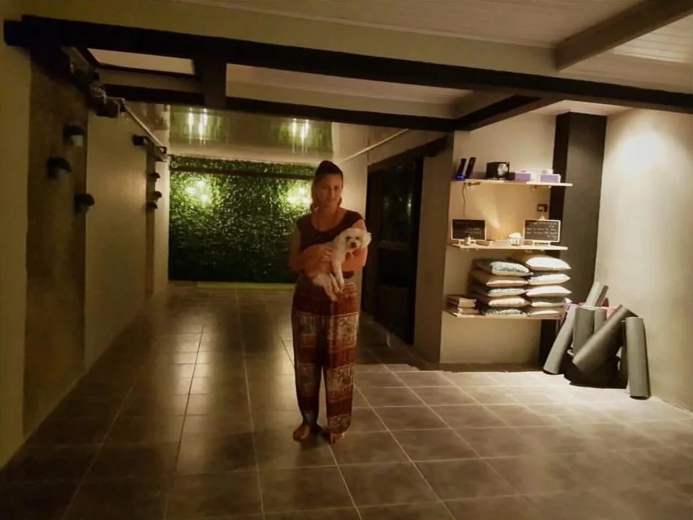 Mujer sosteniendo un perro pequeño en una habitación con paredes decoradas y estantes con libros y objetos.