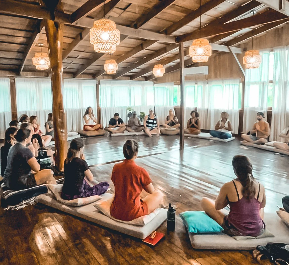 Grupo de personas participando en una clase de meditación o yoga en un salón de madera, sentados en círculo en el suelo y en cojines, en un ambiente calmado y natural con luz suave.