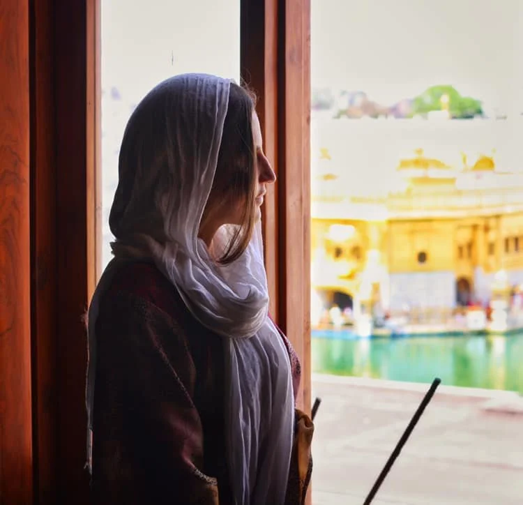 Mujer mirando por una ventana con vista a un cuerpo de agua y construcciones tradicionales.
