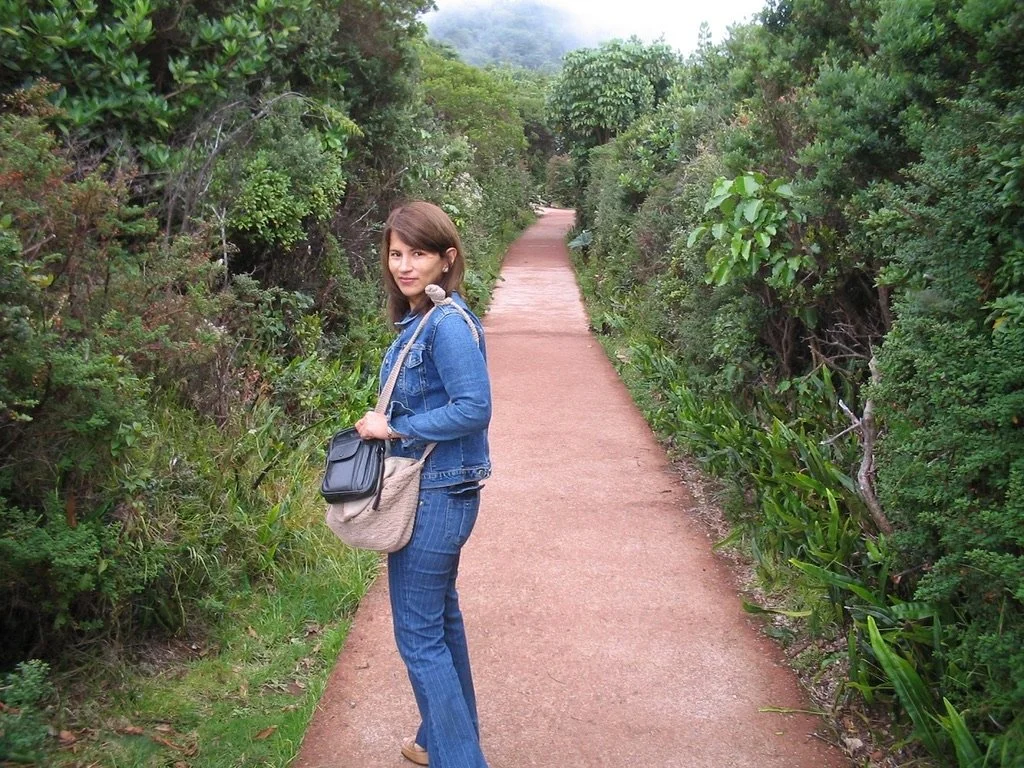 Una mujer con chaqueta de mezclilla y pantalones de mezclilla con una bolsa en la mano, caminando por un sendero rodeado de vegetación densa.