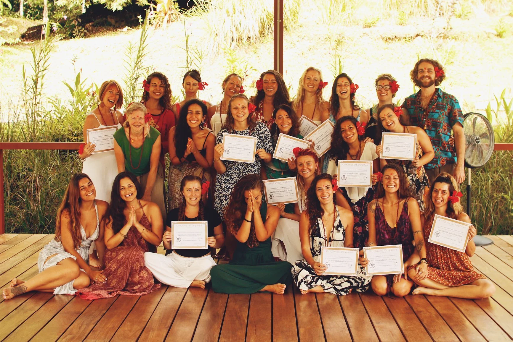 Grupo de personas celebrando, algunas con diplomas y vestimenta colorida, en un ambiente natural con plantas verdes de fondo.