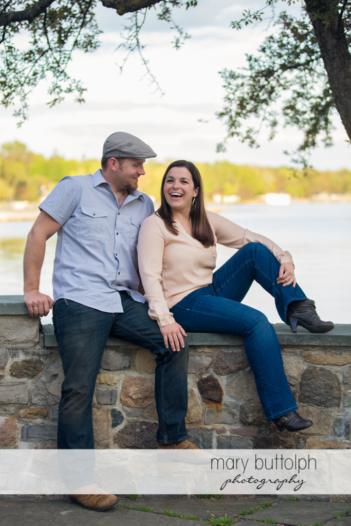 Couple sit on a stone wall near the lake at Skaneateles Engagement