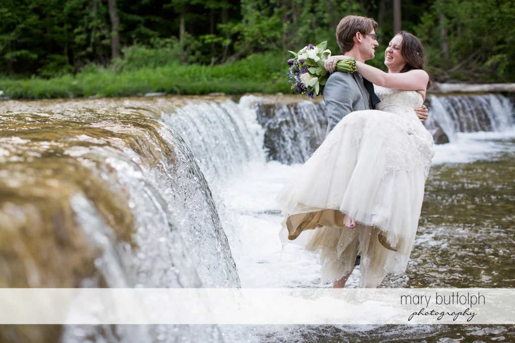 Groom carries the bride across the lake at Taughannock Falls Wedding
