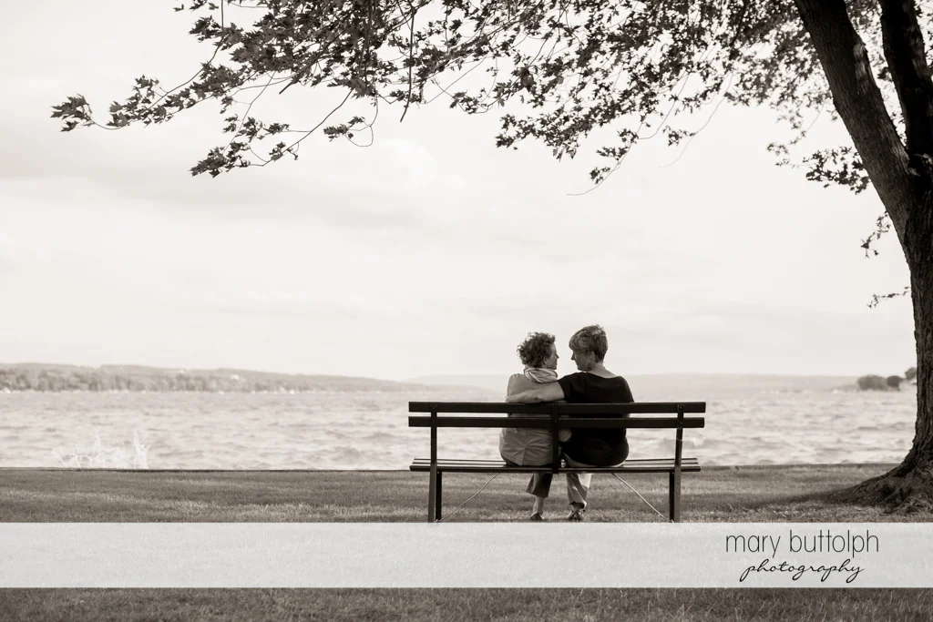 Same sex couple sit on a bench facing the lake at Skaneateles Lake Engagement