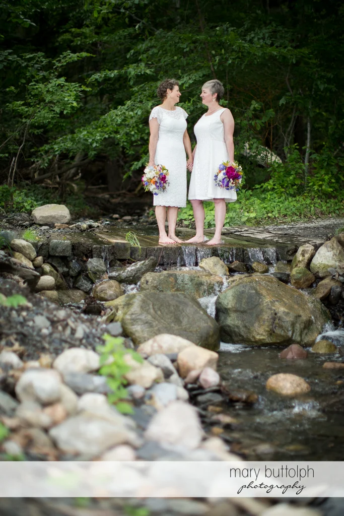 Same sex couple with bouquets pose by the lake at Mandana Barn Wedding