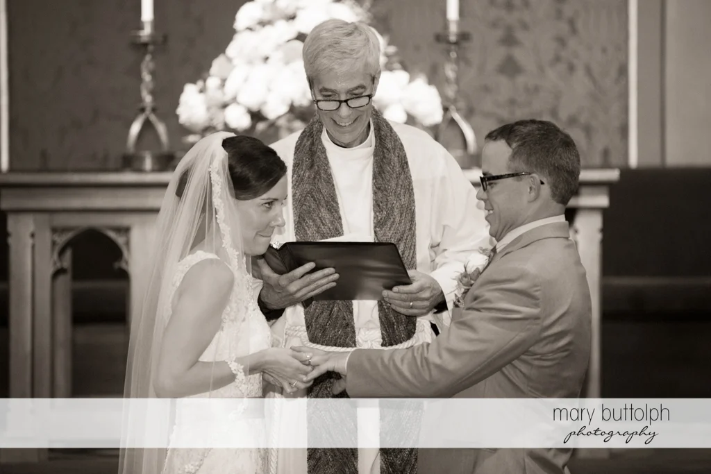 Bride slips the ring on the groom's finger as the priest looks on at the Inns of Aurora Wedding