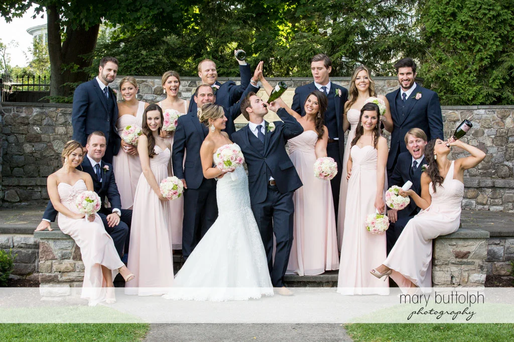 The wedding party pose in the garden as the groom and one of the bridesmaids drink champagne at Skaneateles Country Club Wedding