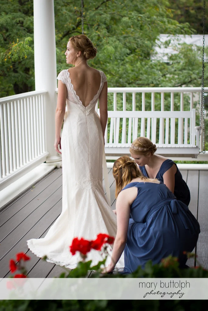 Bride stands on the porch as bridesmaids fix her wedding dress at the Inns of Aurora Wedding