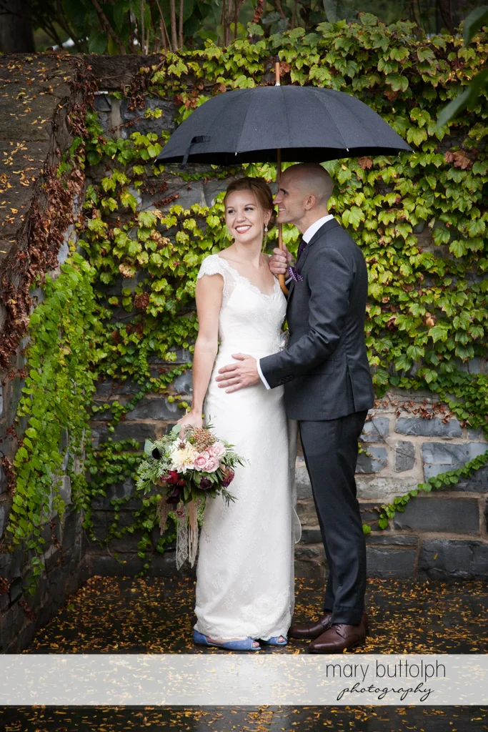 Couple share an umbrella near a brick wall at the Inns of Aurora Wedding