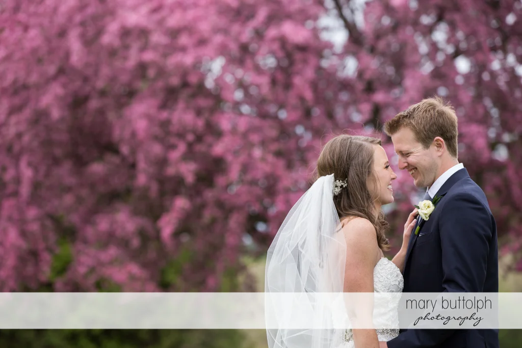 Couple share a happy moment near a tree with purple flowers at Skaneateles Country Club Wedding