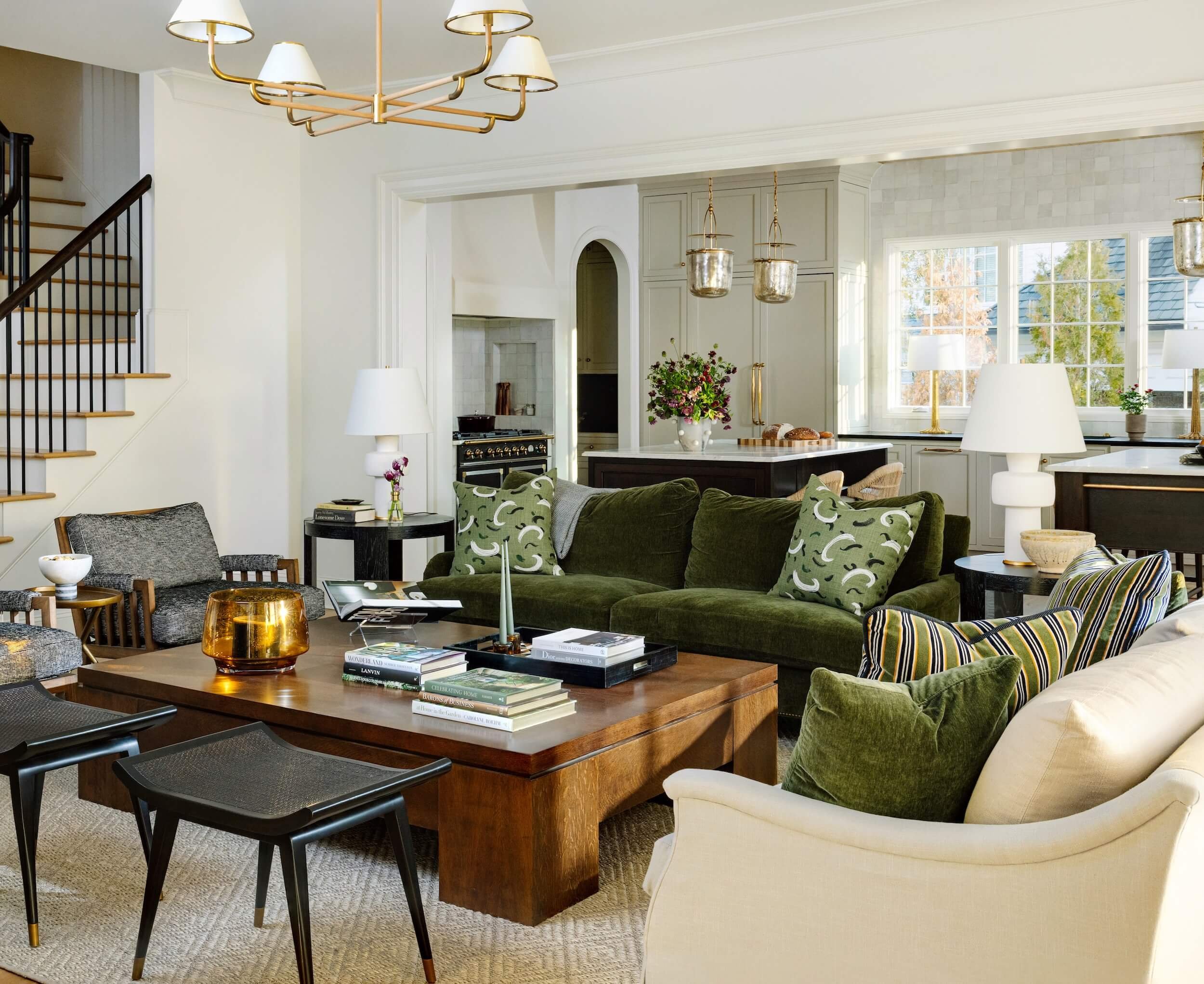 A view of the living room with velvet couch, oversize wooden coffee table, and kitchen behind it