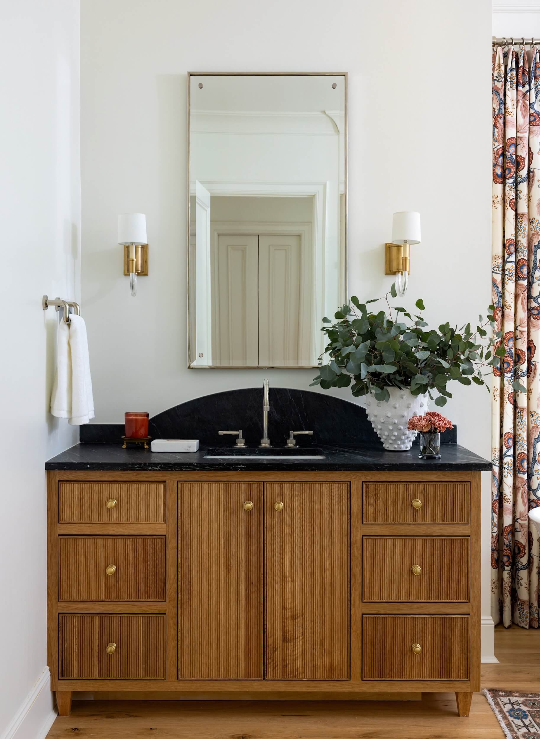 A vanity in the primary bathroom. It is wooden with a dark marble countertop. The mirror above is flanked by 2 sconces
