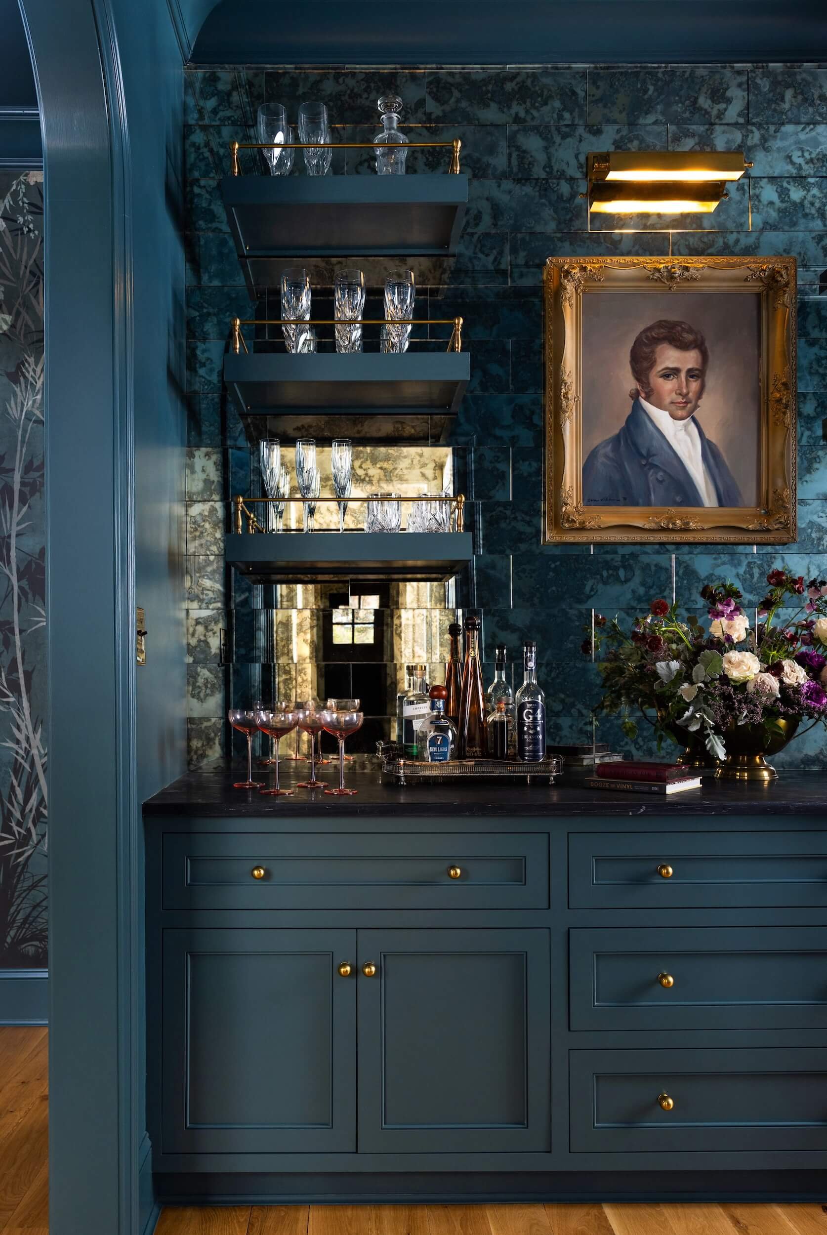 One side of the color drenched butler's pantry with a dark counter top, oil portrait, and covered in glassware