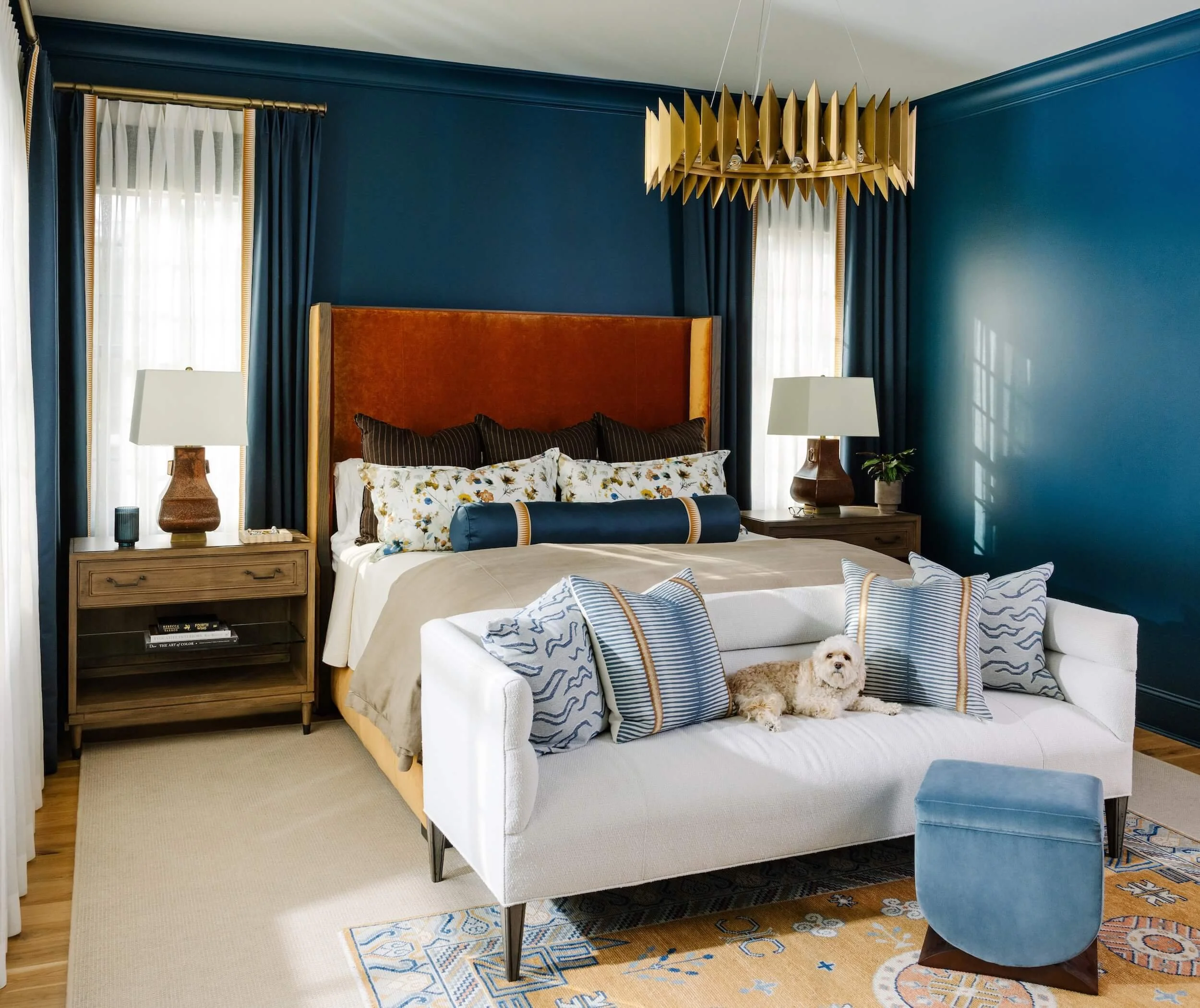 A blue bedroom with velvet headboard and custom pillows. Wooden side tables on either side of the bed and a white couch at the foot of the bed with a small dog sleeping on it