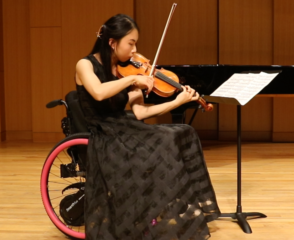 Amy Wang-Hiller seated in a wheelchair performing violin on a recital stage with a grand piano in the background.
