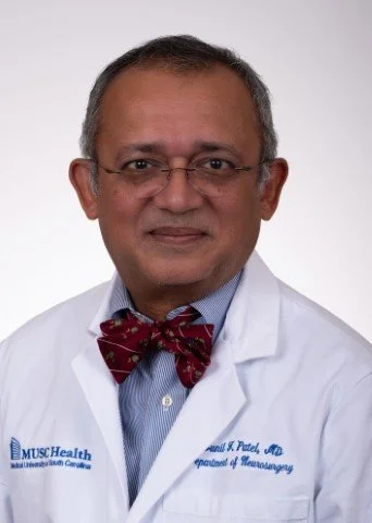 A male healthcare professional in a white lab coat and burgundy bow tie, wearing glasses, standing against a plain background.