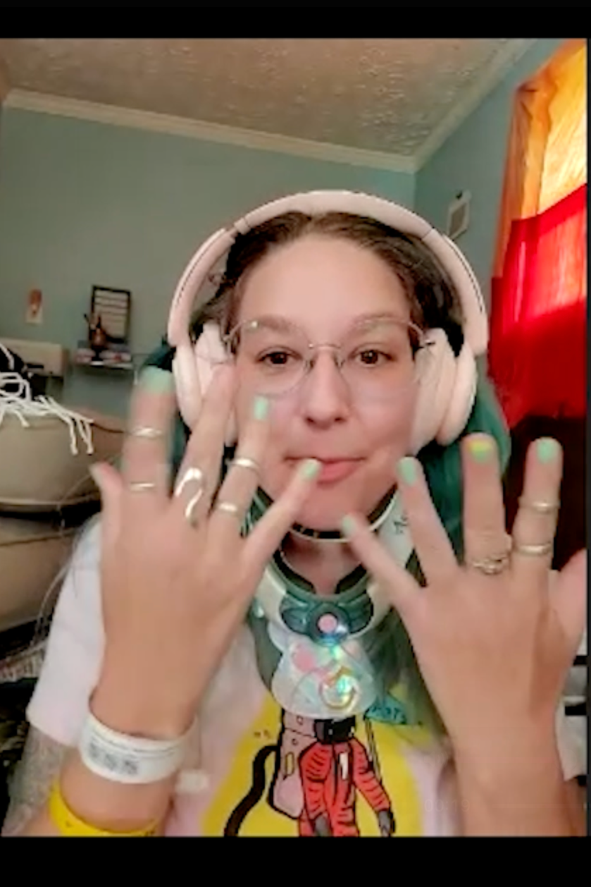 A woman with glasses and headphones showing her hands with multiple silver rings, smiling in a room with colorful curtains and a light blue wall.