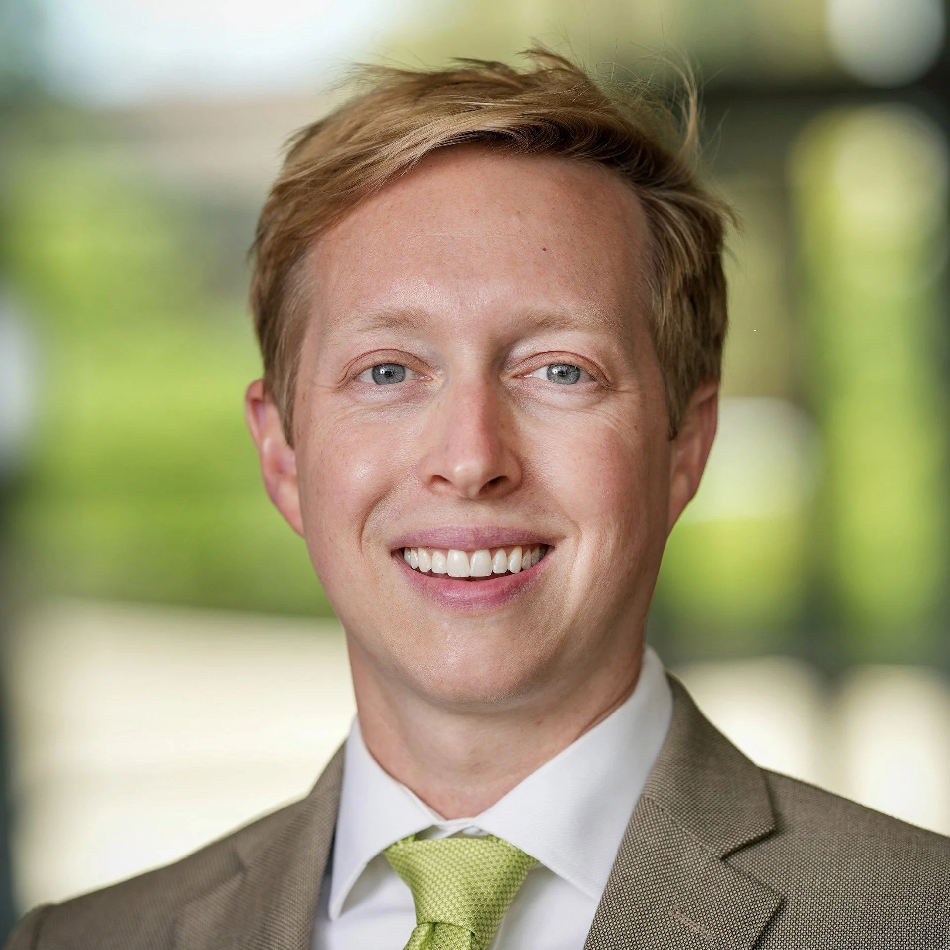 A smiling man with blonde hair, blue eyes, wearing a brown suit, white shirt, and lime green tie, standing outdoors with blurred green foliage in the background.