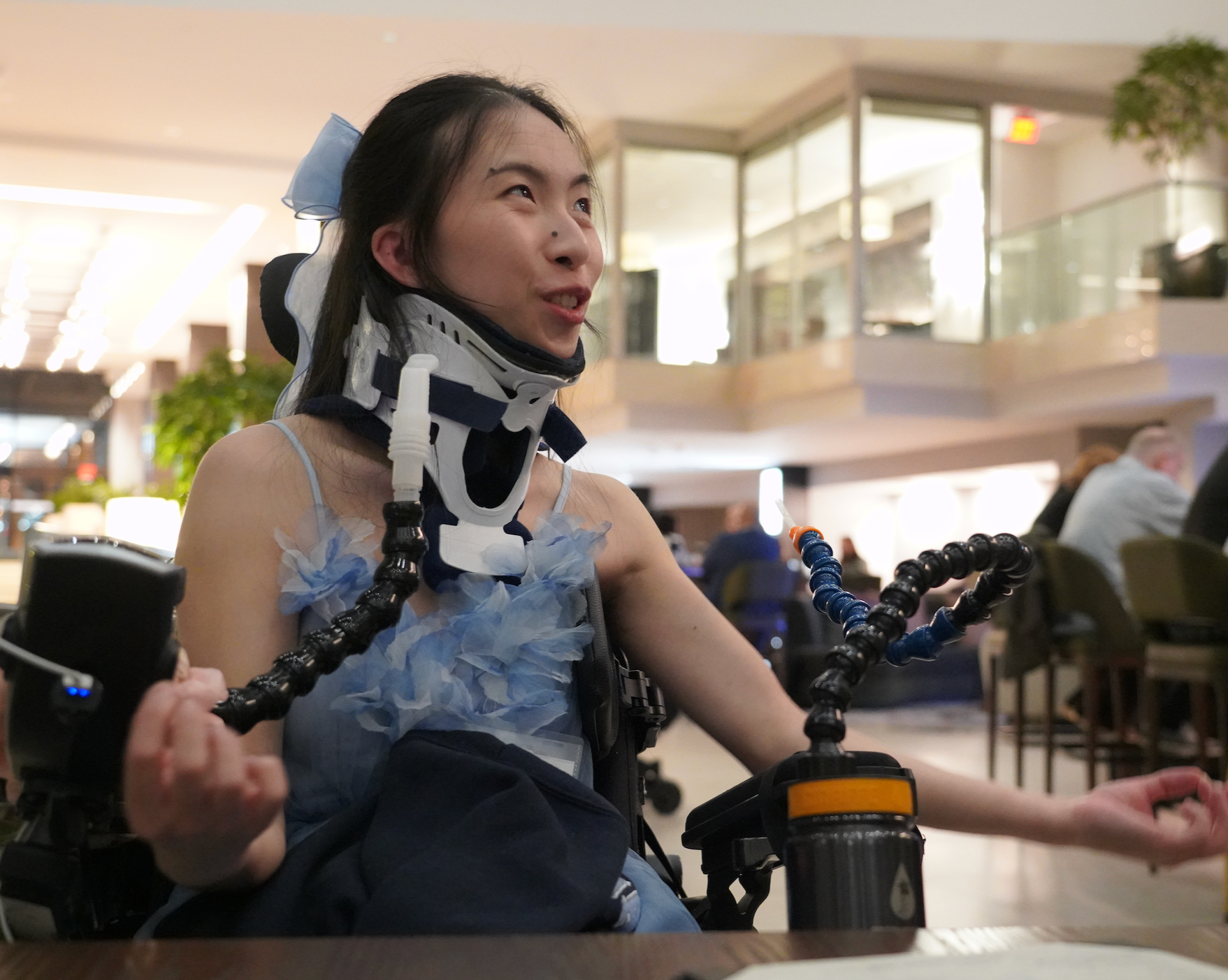 A young woman in a wheelchair with a neck brace and adaptive hand controls, smiling and engaging in conversation in a busy indoor public space.