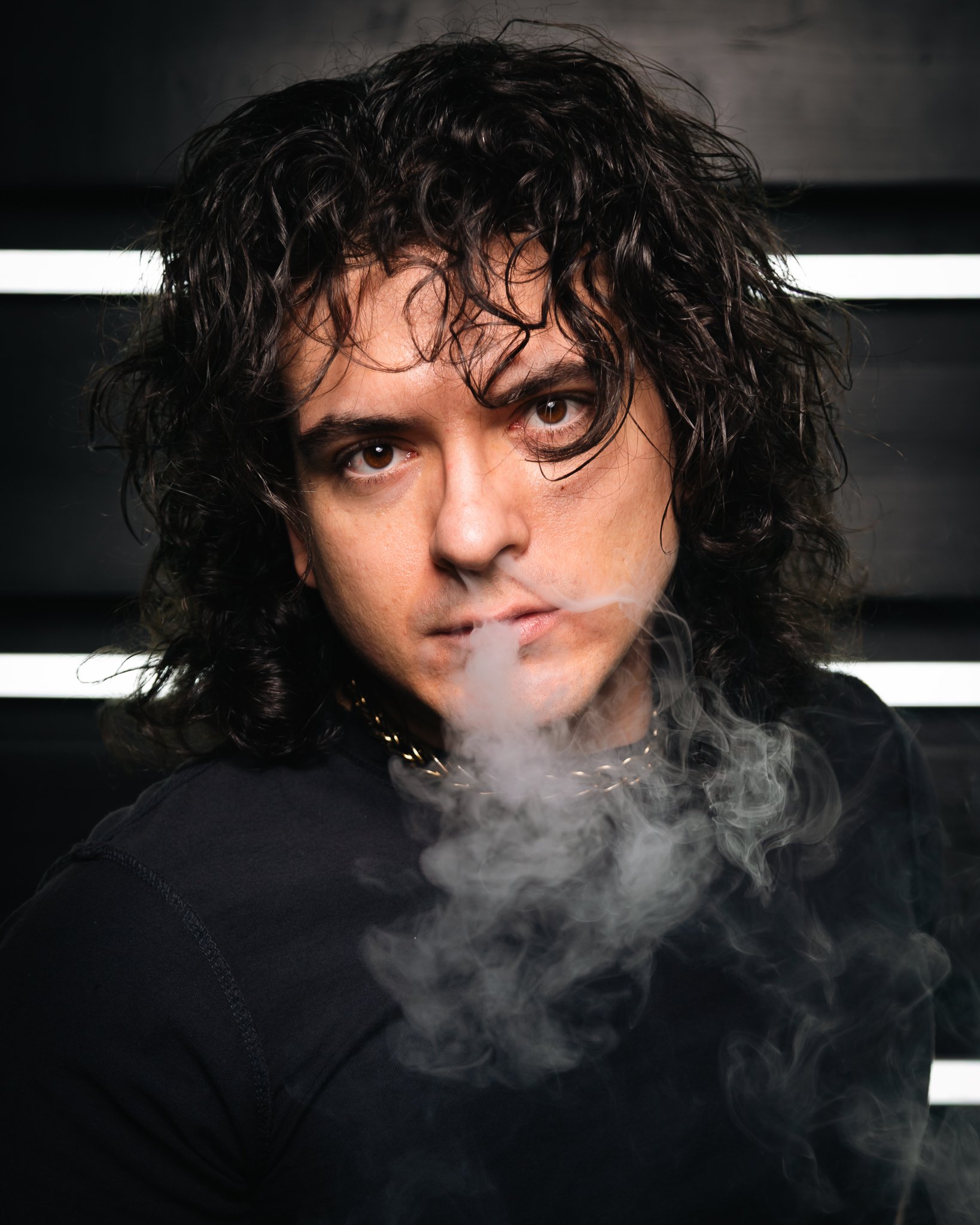 A man with curly dark hair and brown eyes looking directly at the camera, with smoke surrounding his mouth.