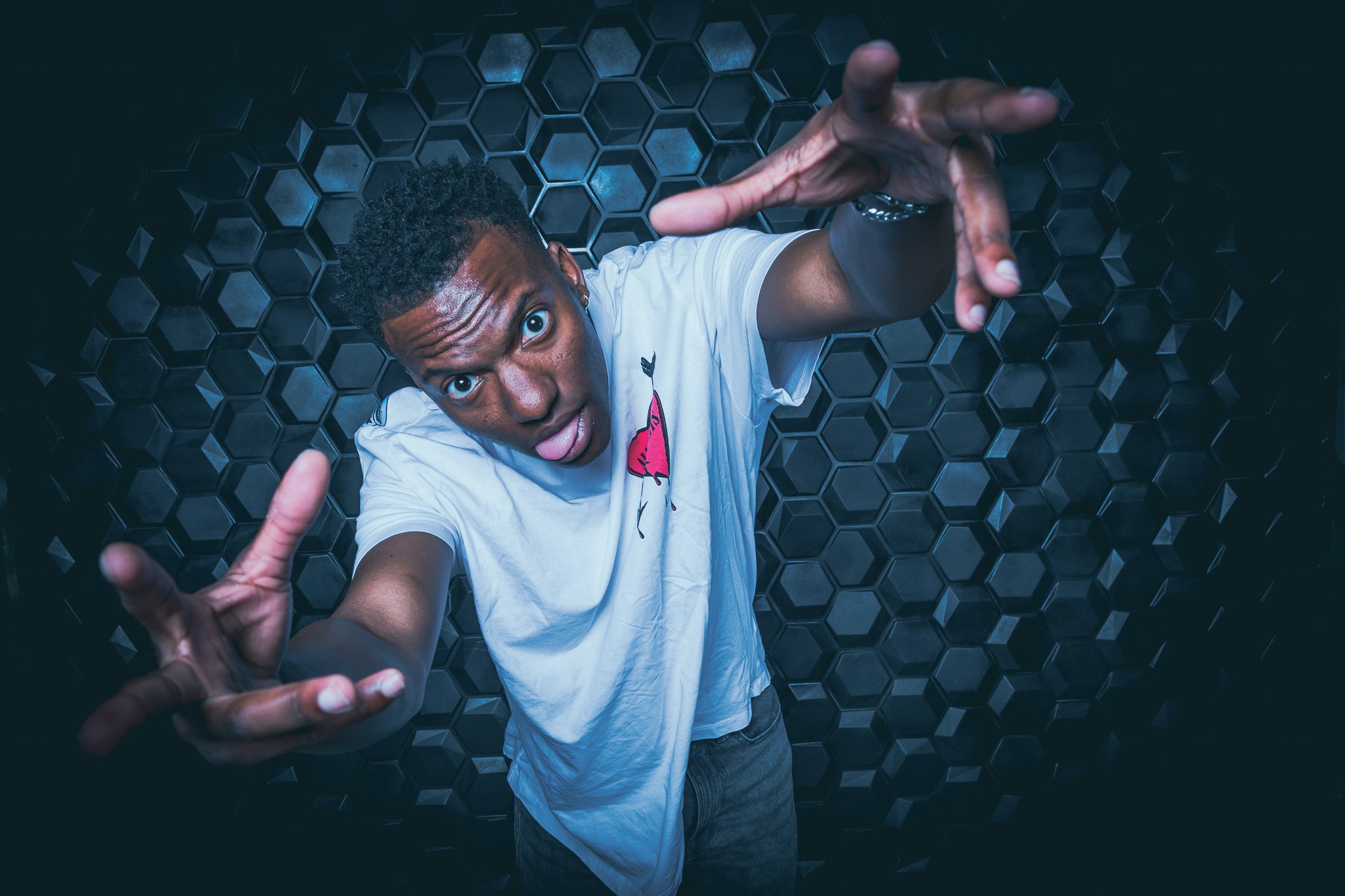 Young man with short curly hair making a reaching gesture towards the camera against a dark honeycomb-patterned background.